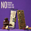 A packaged gluten free millet nut bar stands next to a loose, textured nut bar on a purple background, with stacked chocolate and dried fruit pieces at the base. Large text at the top reads 'NO Maida Palm oil Trans fat'.