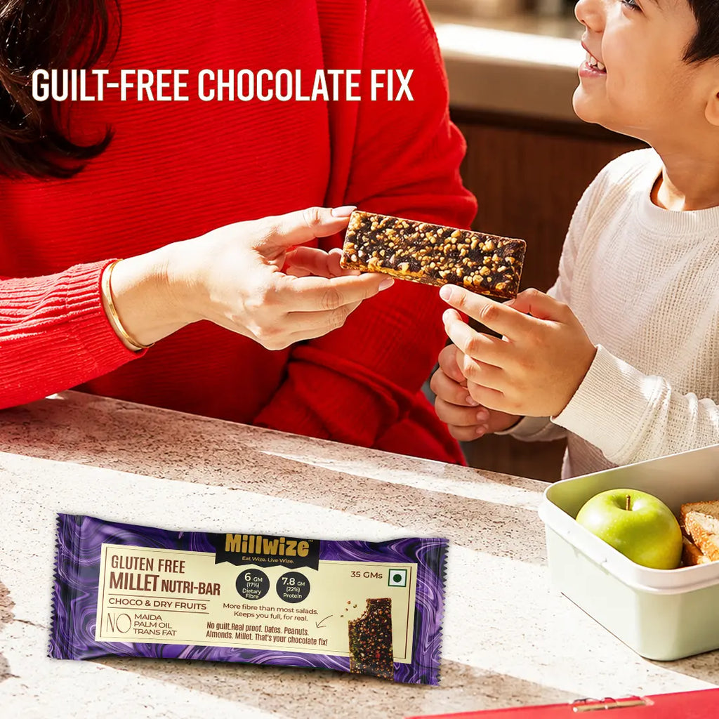 A Millwize gluten free millet nutri-bar is shown being held by an adult and a child in a warm kitchen setting, with the product packaging displayed in the foreground and a green apple and bread in a lunchbox nearby. The text 'GUILT-FREE CHOCOLATE FIX' is displayed at the top left.