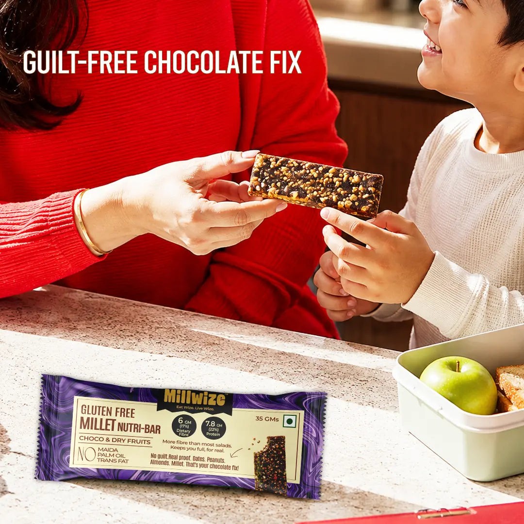 A Millwize gluten free millet nutri-bar is shown being held by an adult and a child in a warm kitchen setting, with the product packaging displayed in the foreground and a green apple and bread in a lunchbox nearby. The text 'GUILT-FREE CHOCOLATE FIX' is displayed at the top left.